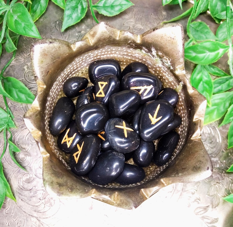 May include: A collection of polished black rune stones with gold-colored symbols, displayed in a decorative silver-colored bowl. The bowl has a unique, petal-like shape. Green foliage surrounds the bowl, adding a natural element to the composition.