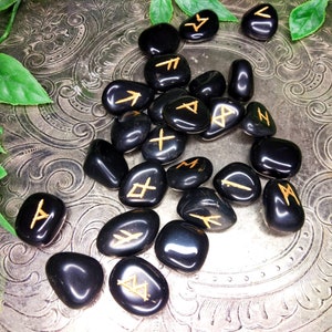 May include: A collection of polished black rune stones with gold-colored symbols. The stones are scattered on a decorative silver surface, with green foliage in the corners. The runes are a form of ancient alphabet used for divination.
