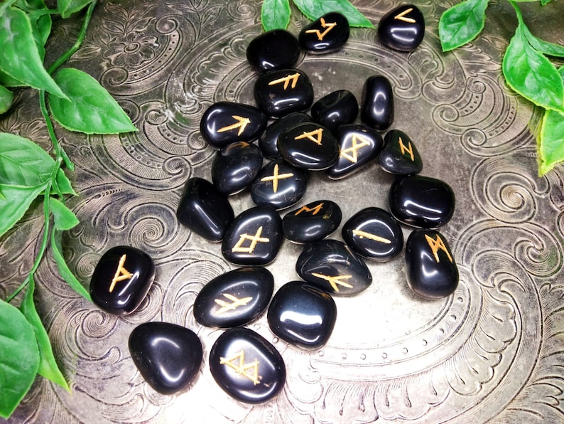 May include: A collection of polished black rune stones with gold-colored symbols. The stones are scattered on a decorative silver surface, with green foliage in the corners. The runes are a form of ancient alphabet used for divination.