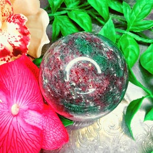 Könnte beinhalten: Eine polierte Edelstein-Kugel mit gesprenkeltem Aussehen, die eine Mischung aus tiefroten und grünen Farbtönen aufweist. Die Kugel wird vor dem Hintergrund von leuchtend rosa und weißen Blumen und grünen Blättern präsentiert, wodurch eine natürliche, dekorative Darstellung entsteht.