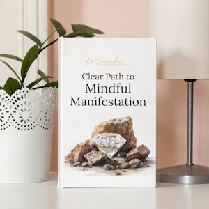May include: A hardcover book titled "Clear Path to Mindful Manifestation" by Dr. Samantha Worthington. The cover features a diamond surrounded by rocks. A white potted plant and a lamp are in the background.