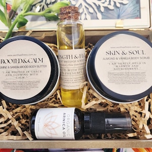 Puede incluir: Una caja de regalo de madera llena de productos para el cuidado personal. Incluye manteca corporal, exfoliante corporal, aceite de masaje y aceite de árnica. Los productos están en frascos y botellas con etiquetas.