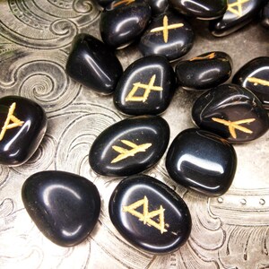 May include: A collection of polished black rune stones with gold-colored symbols. The stones are scattered on a silver, textured surface. The runes are in various shapes and sizes, with a glossy finish.