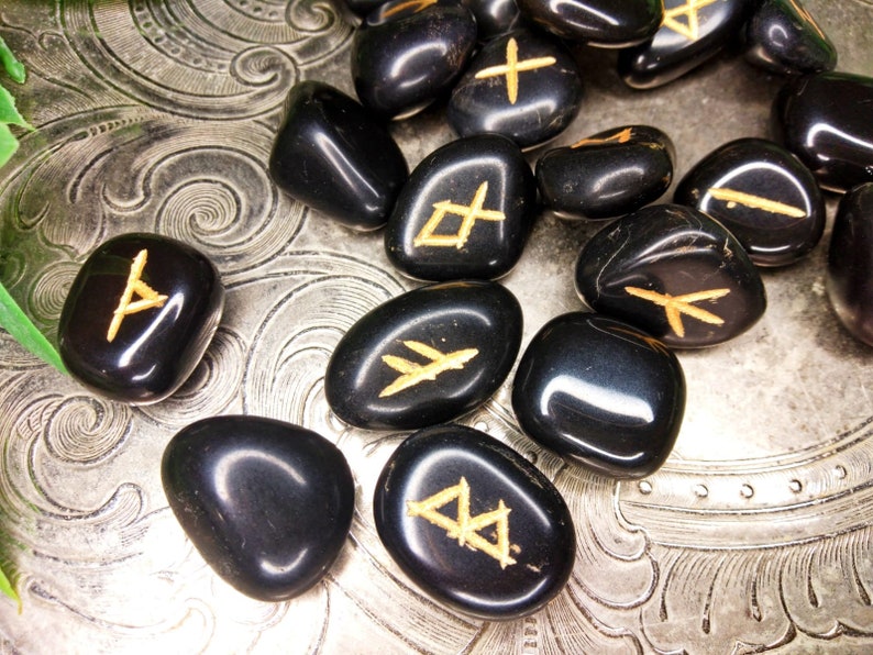 May include: A collection of polished black rune stones with gold-colored symbols. The stones are scattered on a silver, textured surface. The runes are in various shapes and sizes, with a glossy finish.