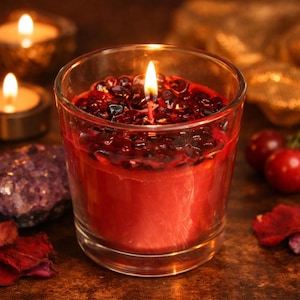 Puede incluir: Una vela roja encendida en un vaso transparente. La vela está adornada con piedras de color rojo oscuro y negro. Otras velas encendidas y elementos decorativos están en el fondo. La vela está sobre una superficie marrón.
