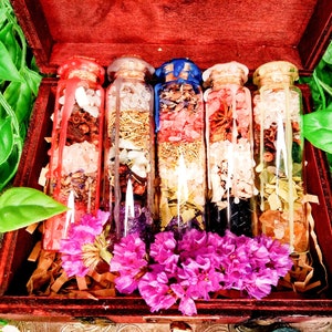 Könnte beinhalten: Eine dunkle Holzkiste enthält fünf Glasflaschen, gefüllt mit bunten getrockneten Kräutern und Blumen. Die Flaschen sind mit Korken und Wachs verschlossen. Rosa Blüten und braune Papierschnipsel schmücken die Kiste.