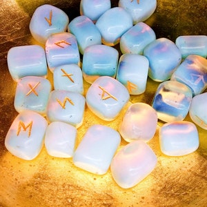 May include: A collection of opalescent rune stones with gold-colored symbols. The stones are a mix of pale blue and white, with a soft, glowing appearance. They are arranged in a golden bowl, creating a mystical and ethereal aesthetic.