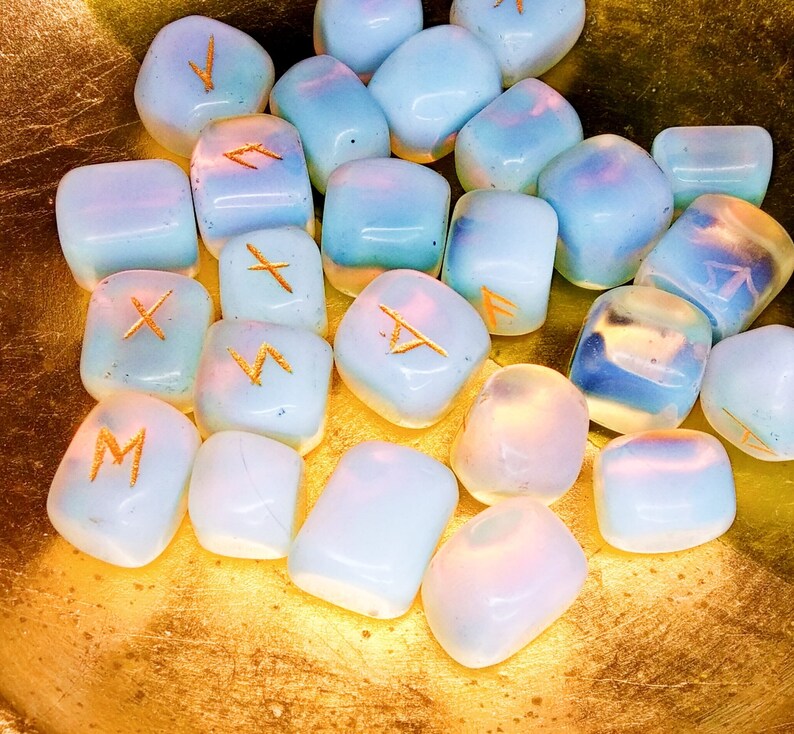May include: A collection of opalescent rune stones with gold-colored symbols. The stones are a mix of pale blue and white, with a soft, glowing appearance. They are arranged in a golden bowl, creating a mystical and ethereal aesthetic.