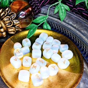 May include: A collection of opalescent rune stones with gold-colored symbols, arranged on a gold-colored plate. The stones are smooth and rounded, with a soft, glowing appearance. The background includes decorative elements and greenery.
