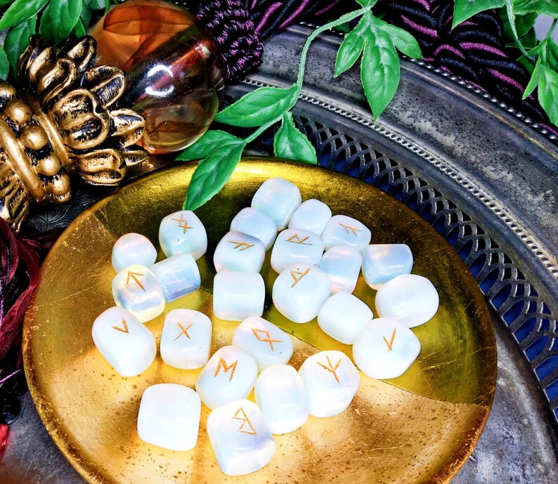 May include: A collection of opalescent rune stones with gold-colored symbols, arranged on a gold-colored plate. The stones are smooth and rounded, with a soft, glowing appearance. The background includes decorative elements and greenery.