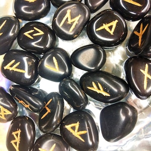 May include: A collection of polished black stones, each engraved with a golden rune symbol. The stones vary in size and shape, with a glossy finish. The runes are in a golden color, contrasting against the dark background.