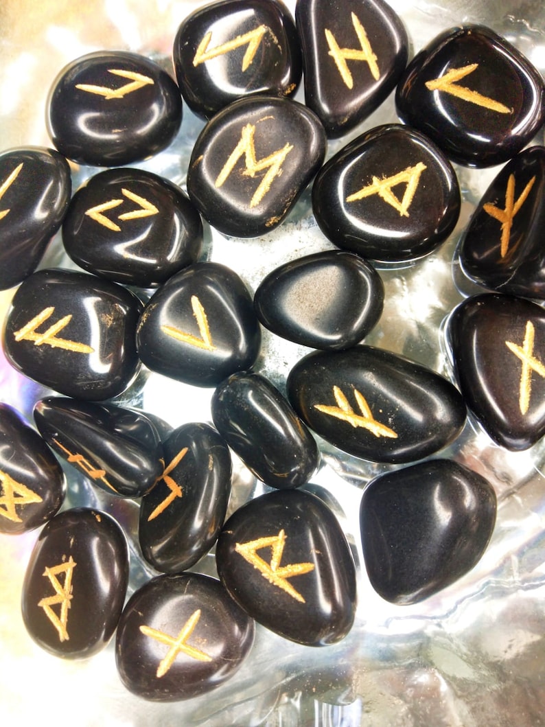 May include: A collection of polished black stones, each engraved with a golden rune symbol. The stones vary in size and shape, with a glossy finish. The runes are in a golden color, contrasting against the dark background.