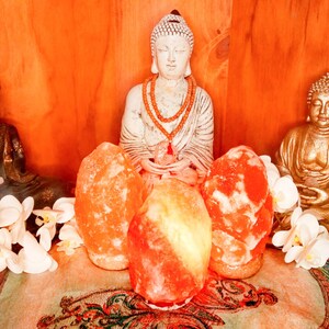 以下が含まれることがあります： 黒、白、金色の3体の仏像と、3つのピンク色のヒマラヤ岩塩ランプが並んだ穏やかなディスプレイ。白い蘭と模様のある布が、静かな雰囲気を添えています。