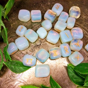 May include: A collection of opalescent rune stones with gold-colored symbols, arranged on a decorative silver tray with green foliage. The stones are smooth and rounded, with a soft, ethereal glow. The runes are etched into the surface.