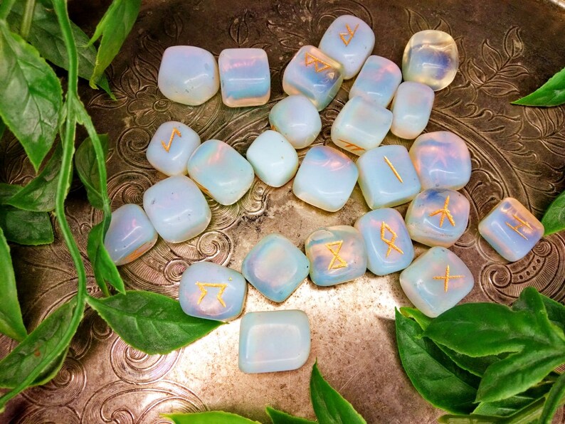 May include: A collection of opalescent rune stones with gold-colored symbols, arranged on a decorative silver tray with green foliage. The stones are smooth and rounded, with a soft, ethereal glow. The runes are etched into the surface.
