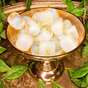 May include: A collection of opalescent rune stones with gold symbols rests in a gold-colored bowl. The bowl sits on a decorative silver tray, surrounded by green leafy vines. The runes are smooth and rounded.