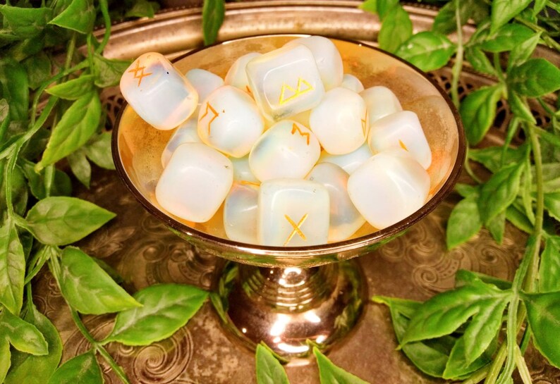 May include: A collection of opalescent rune stones with gold symbols rests in a gold-colored bowl. The bowl sits on a decorative silver tray, surrounded by green leafy vines. The runes are smooth and rounded.