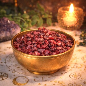 May include: A brass bowl overflowing with small, irregularly shaped, deep red and purple gemstones. The bowl is on a patterned surface, with a lit candle and a purple crystal in the background. The gemstones appear tumbled.