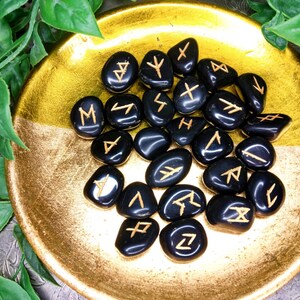 May include: A collection of black rune stones with gold-colored symbols, displayed in a gold-colored bowl. The stones are smooth and polished, with each bearing a unique symbol. The bowl has a textured surface and is surrounded by green foliage.