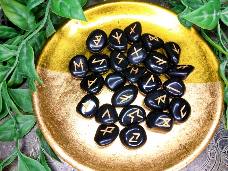 May include: A collection of black rune stones with gold-colored symbols, displayed in a gold-colored bowl. The stones are smooth and polished, with each bearing a unique symbol. The bowl has a textured surface and is surrounded by green foliage.