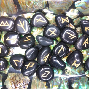 May include: A collection of polished black rune stones with gold-colored symbols. The stones are smooth and oval-shaped, resting on a surface with iridescent shell fragments. The runes are clearly etched.