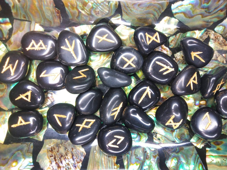 May include: A collection of polished black rune stones with gold-colored symbols. The stones are smooth and oval-shaped, resting on a surface with iridescent shell fragments. The runes are clearly etched.