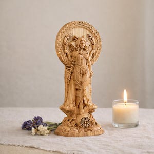 May include: A carved wooden statue of a goddess figure, holding torches and standing before a circular wreath. The statue is on a base with a circular symbol. A lit candle in a glass holder and dried flowers are in the foreground.