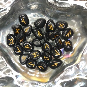 May include: A collection of black, polished rune stones with gold-colored symbols. The stones are arranged in a silver, iridescent bowl. The runes are a form of ancient Germanic alphabet, used for divination and spiritual practices.