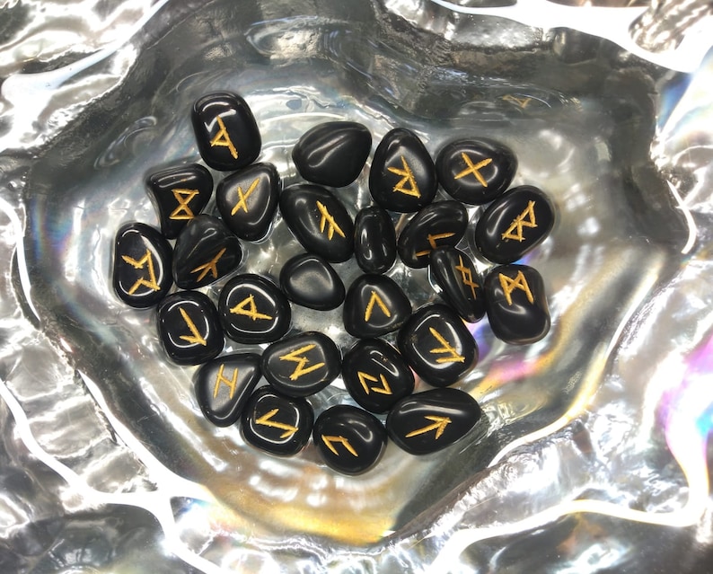 May include: A collection of black, polished rune stones with gold-colored symbols. The stones are arranged in a silver, iridescent bowl. The runes are a form of ancient Germanic alphabet, used for divination and spiritual practices.