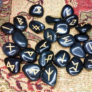 May include: A collection of black, polished rune stones with gold-colored etched symbols. The stones are scattered on a patterned, multi-colored fabric. The runes are a divination tool, used for spiritual guidance and insight.