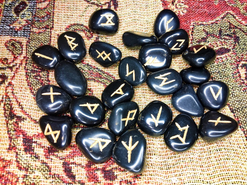 May include: A collection of black, polished rune stones with gold-colored etched symbols. The stones are scattered on a patterned, multi-colored fabric. The runes are a divination tool, used for spiritual guidance and insight.
