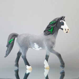 May include: A gray horse figurine with a white blaze on its face, white markings on its lower legs, and a white patch on its belly. The horse has a dark gray mane and tail accented with bright green strands and dark gray lower legs.