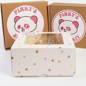 May include: Two brown cardboard boxes featuring a circular sticker with a pink and white panda illustration and the text Pinky's Slime Kit. In front sits a white box with a clear window and colorful polka dots with gold foil accents.