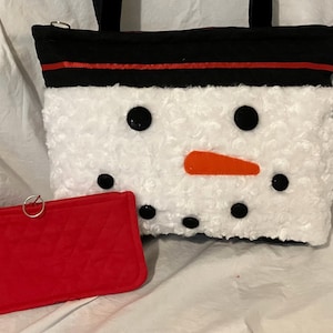 May include: A white, fluffy snowman-themed tote bag with black eyes and mouth, and an orange nose. The bag has a black top with a red stripe and black handles. A small red wallet with a silver ring is in front.