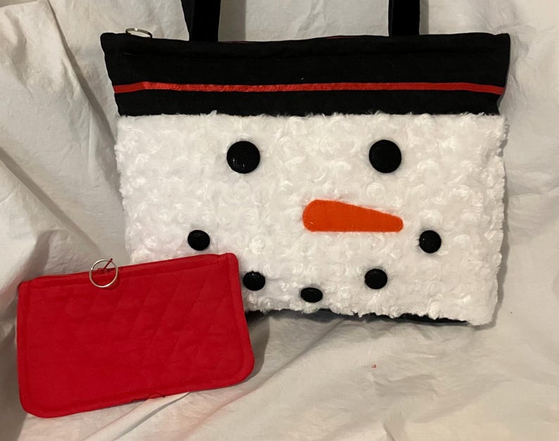 May include: A white, fluffy snowman-themed tote bag with black eyes and mouth, and an orange nose. The bag has a black top with a red stripe and black handles. A small red wallet with a silver ring is in front.