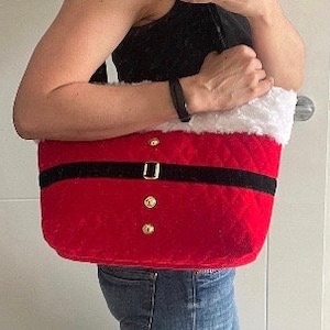 May include: Red Santa Claus-themed tote bag with a black belt and gold button details. The top edge is trimmed with white faux fur. The bag is held by a person wearing a black top and blue jeans.