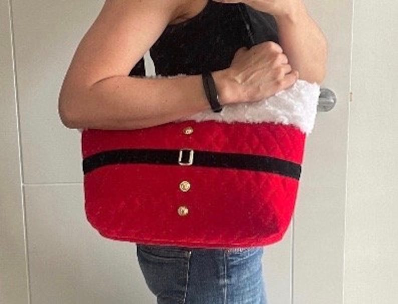May include: Red Santa Claus-themed tote bag with a black belt and gold button details. The top edge is trimmed with white faux fur. The bag is held by a person wearing a black top and blue jeans.