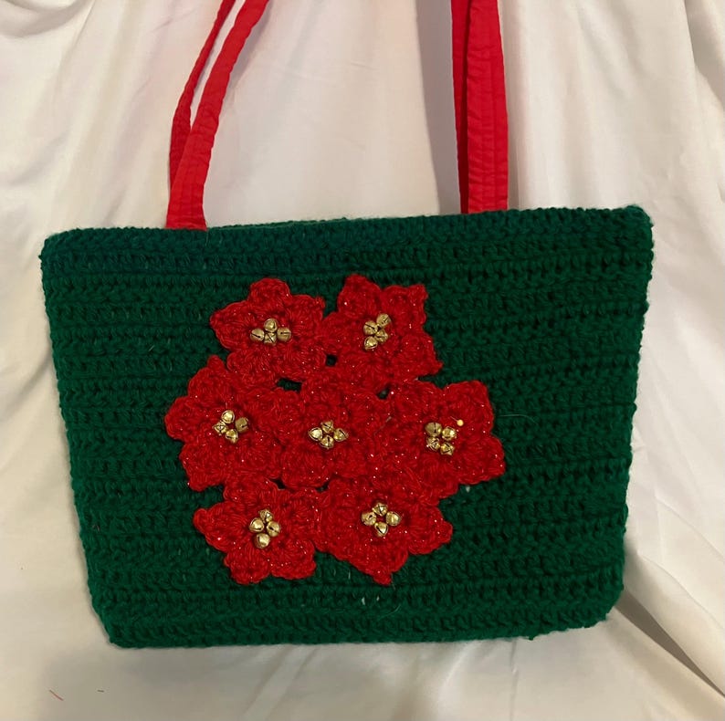 May include: A handmade green crochet tote bag with red flower embellishments and red handles. The flowers have gold-colored bead centers. The bag is rectangular and appears to be made of yarn.
