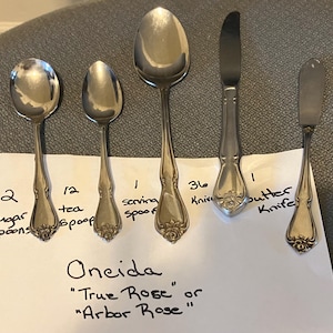 May include: A set of silver-toned stainless steel flatware including sugar spoons, tea spoons, a serving spoon, knives, and a butter knife, all featuring a rose motif on the handle. A paper label lists the Oneida True Rose or Arbor Rose pattern.