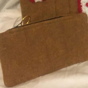May include: A brown rectangular zippered pouch with a gold-colored ring on the zipper pull. The pouch is made of a soft, textured material. In the background, a portion of a gingerbread-themed item is visible.
