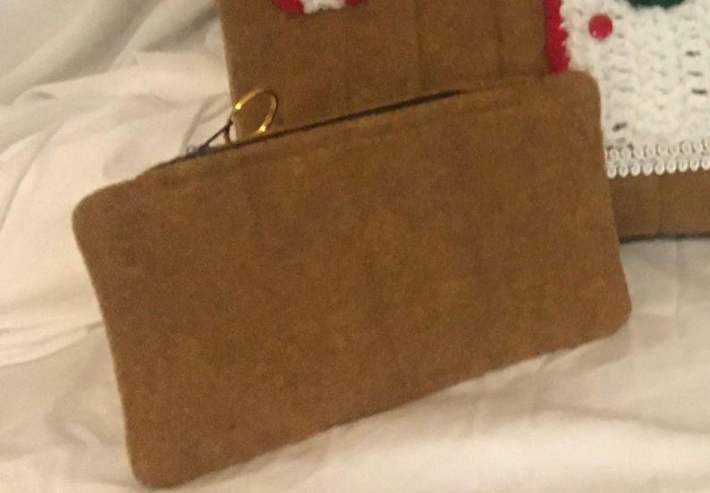 May include: A brown rectangular zippered pouch with a gold-colored ring on the zipper pull. The pouch is made of a soft, textured material. In the background, a portion of a gingerbread-themed item is visible.