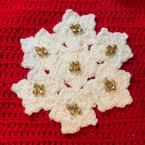 May include: A white crocheted snowflake with gold-colored embellishments, set against a red crocheted background. The snowflake is made of seven flower-shaped elements, each adorned with a cluster of small, gold-colored beads.