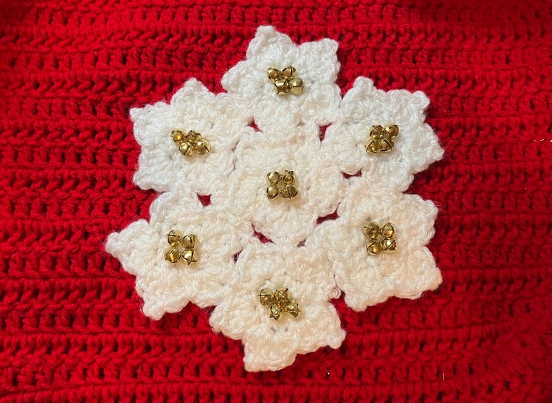 May include: A white crocheted snowflake with gold-colored embellishments, set against a red crocheted background. The snowflake is made of seven flower-shaped elements, each adorned with a cluster of small, gold-colored beads.