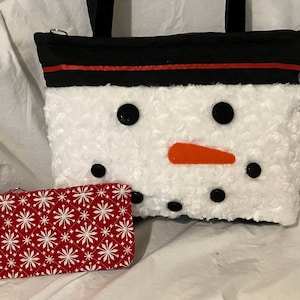 May include: A white snowman-themed tote bag with black button eyes, an orange carrot nose, and a black top with a red stripe. A red and white snowflake-patterned wallet is next to the bag.