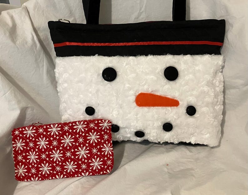 May include: A white snowman-themed tote bag with black button eyes, an orange carrot nose, and a black top with a red stripe. A red and white snowflake-patterned wallet is next to the bag.