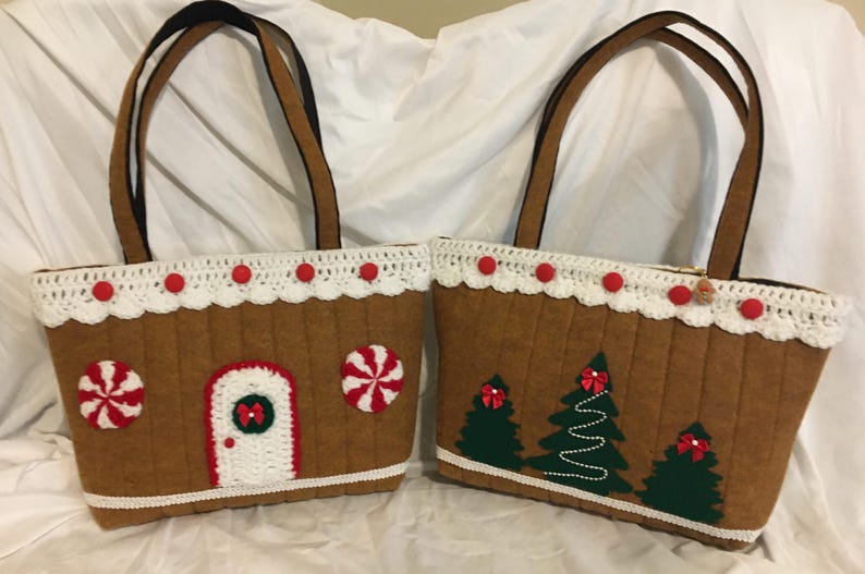 May include: Two brown tote bags with gingerbread house designs. Each bag features a white crocheted trim, red accents, and a black handle. One bag has a door and candy accents, the other has Christmas tree designs.