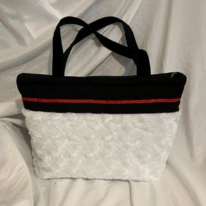May include: A black tote bag with a white, textured bottom and black handles. A thin red stripe accents the top of the white section. The bag is sitting on a white surface.