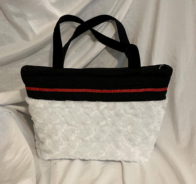 May include: A black tote bag with a white, textured bottom and black handles. A thin red stripe accents the top of the white section. The bag is sitting on a white surface.
