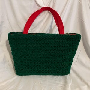 May include: A handmade green crochet tote bag with red handles. The bag has a rectangular shape with a red interior lining. The handles are made of a soft, velvety material. The bag is displayed on a white background.