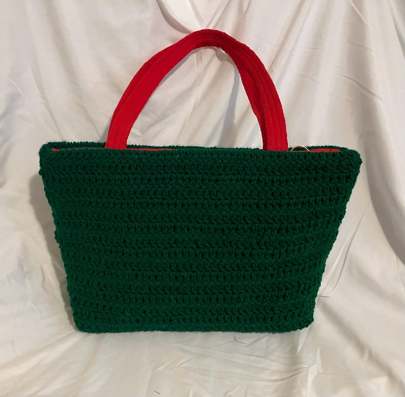 May include: A handmade green crochet tote bag with red handles. The bag has a rectangular shape with a red interior lining. The handles are made of a soft, velvety material. The bag is displayed on a white background.
